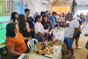 Imagem colorida mostra estudantes apresentando vinho de açaí para visitantes do Sairé 2025