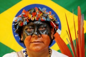 Imagem colorida mostra uma foto de um rosto de uma mulher indígena com rosto pintado e bandeira do Brasil ao fundo para representar os povos indígenas