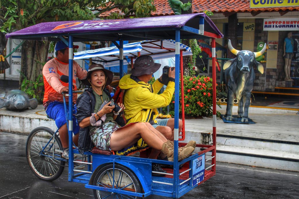 Imagem colorida mostra duas pessoas em triciclo de Parintins fotografando pontos da cidade de Parintins