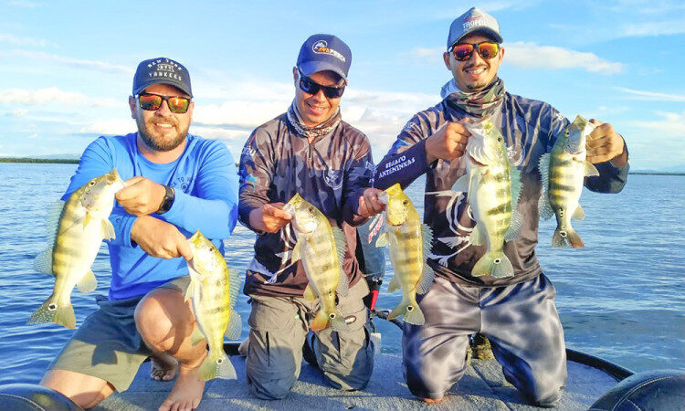 Imagem colorida mostra e homens em uma atividade de pesca esportiva segurando peixes na beira de um rio em tocantins
