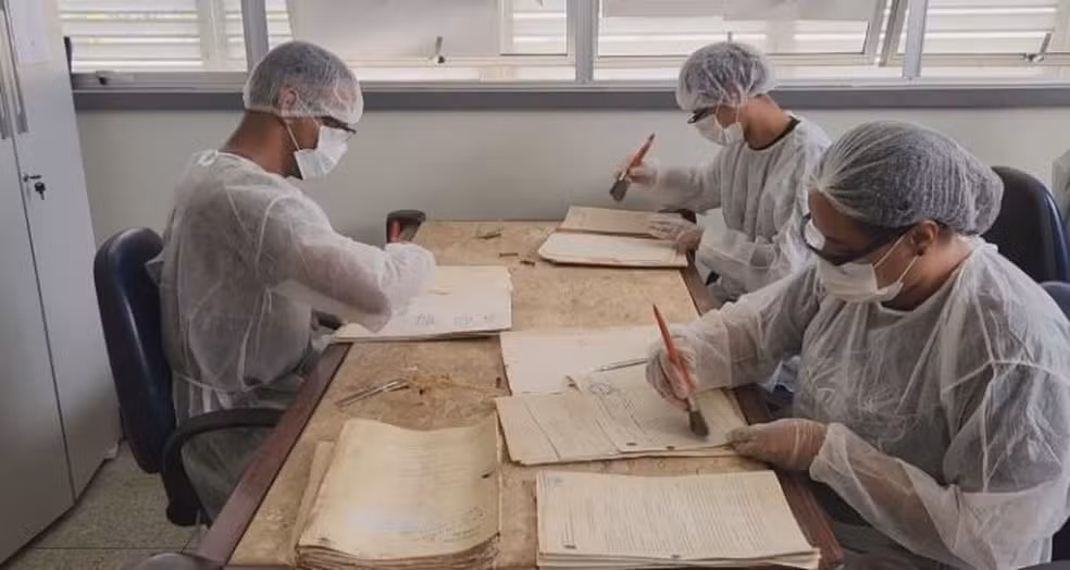Imagem colorida mostra três pessoas fazendo trabalhos de recuperação de documentos antigos