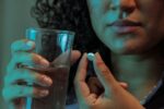 Imagem colorida mostra mulher segurando copo com água e remédio em outra mão realizando automedicação