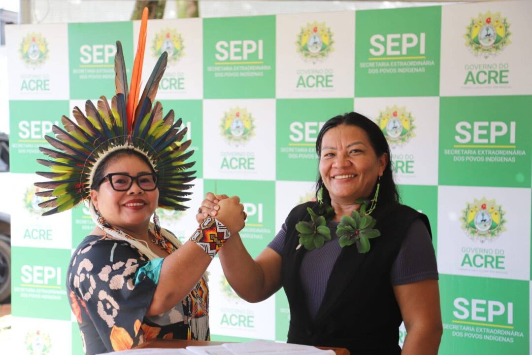 Imagem colorida mostra duas mulheres indígenas se cumprimentando e olhando para foto sorrindo