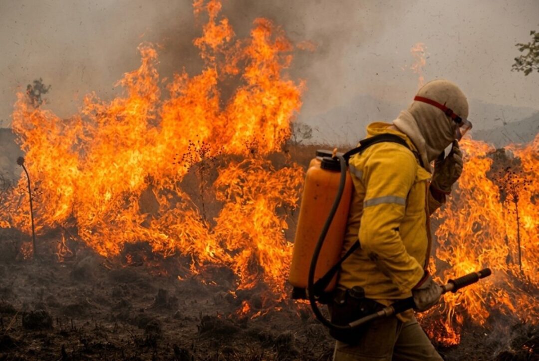 Imagem colorida mostra homeme equipado para combate a incêndio e chamas ao fundo que provocam fumaças