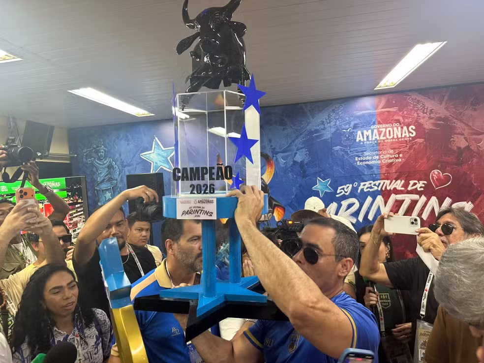 Imagem colorida mostra presidente do Caprichoso Rossi Amoêdo segurando troféu de campeão, cercado de pessoas em espaço pequeno fechado
