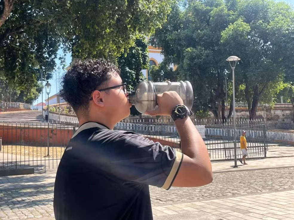 Imagem colorida mostra homem bebendo água em garrafa de armazenamento sob forte calor em uma praça de Manaus